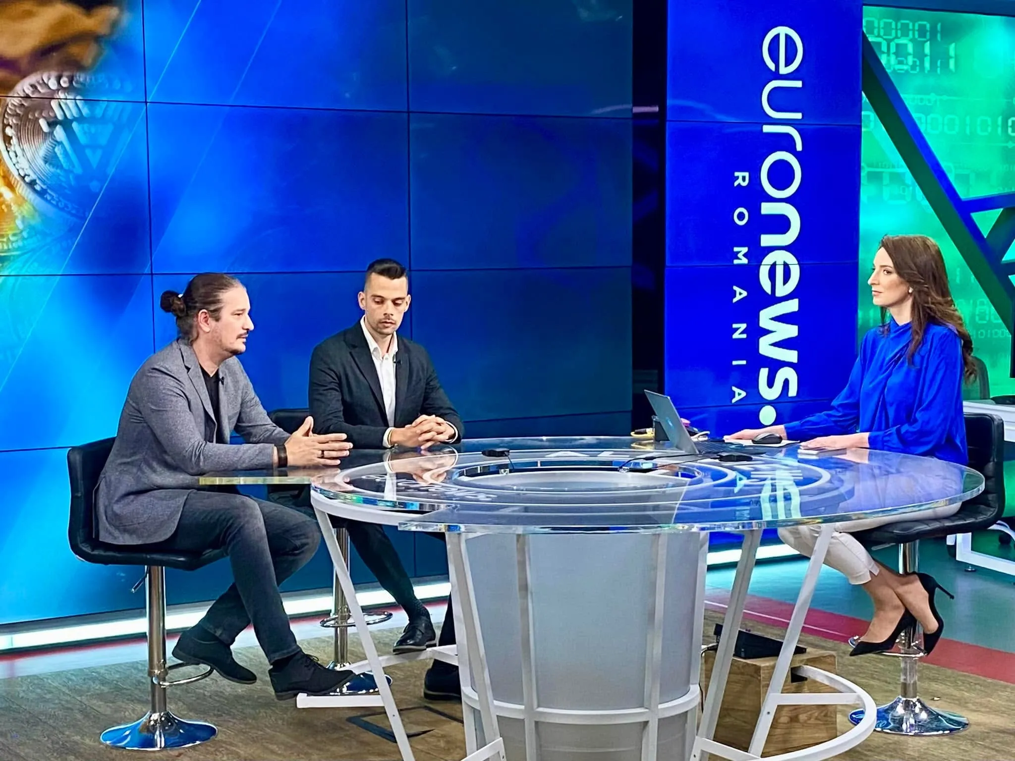 Three people sit at a transparent round table in the Euronews Romania studio during a broadcast.