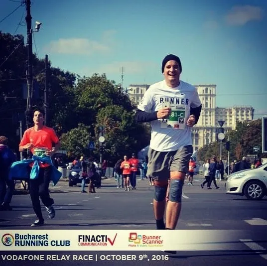 A smiling runner in a white RUNNER shirt, black beanie, and race bib jogs past other participants with Bucharest’s Palace of the Parliament behind him