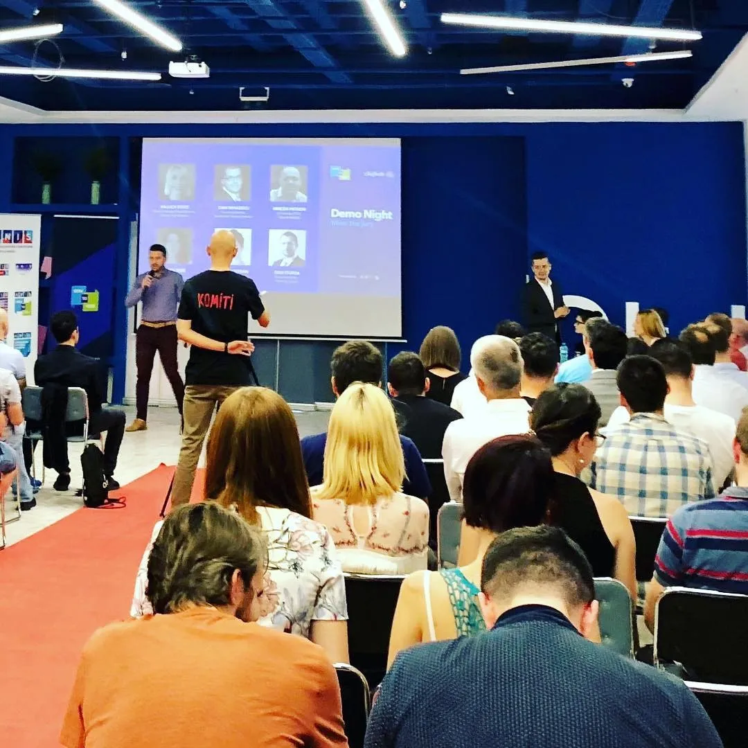 Audience watches presenters on stage under a Demo Night slide at an indoor event with blue walls and a red carpet.
