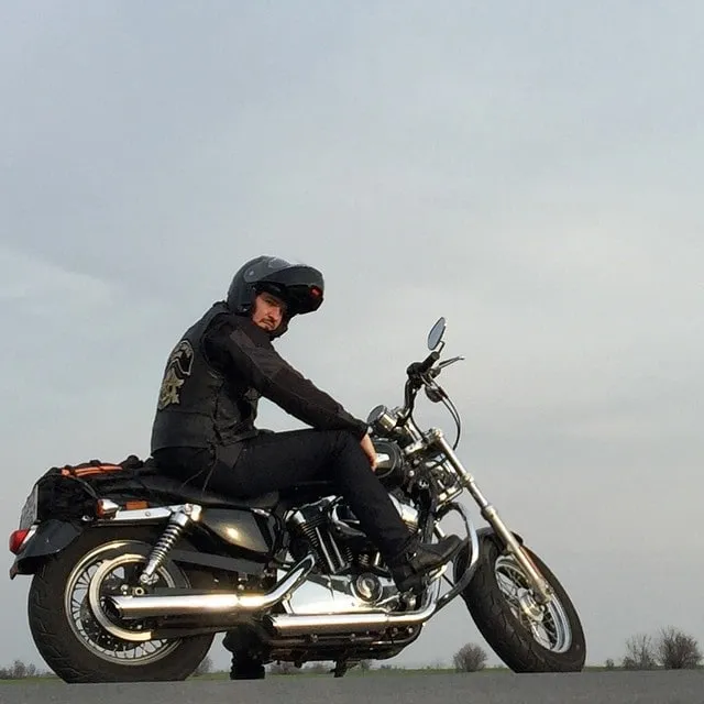 Un bărbat cu o cască neagră și o vestă de piele stă pe o motocicletă, pe fundalul unui cer pal.