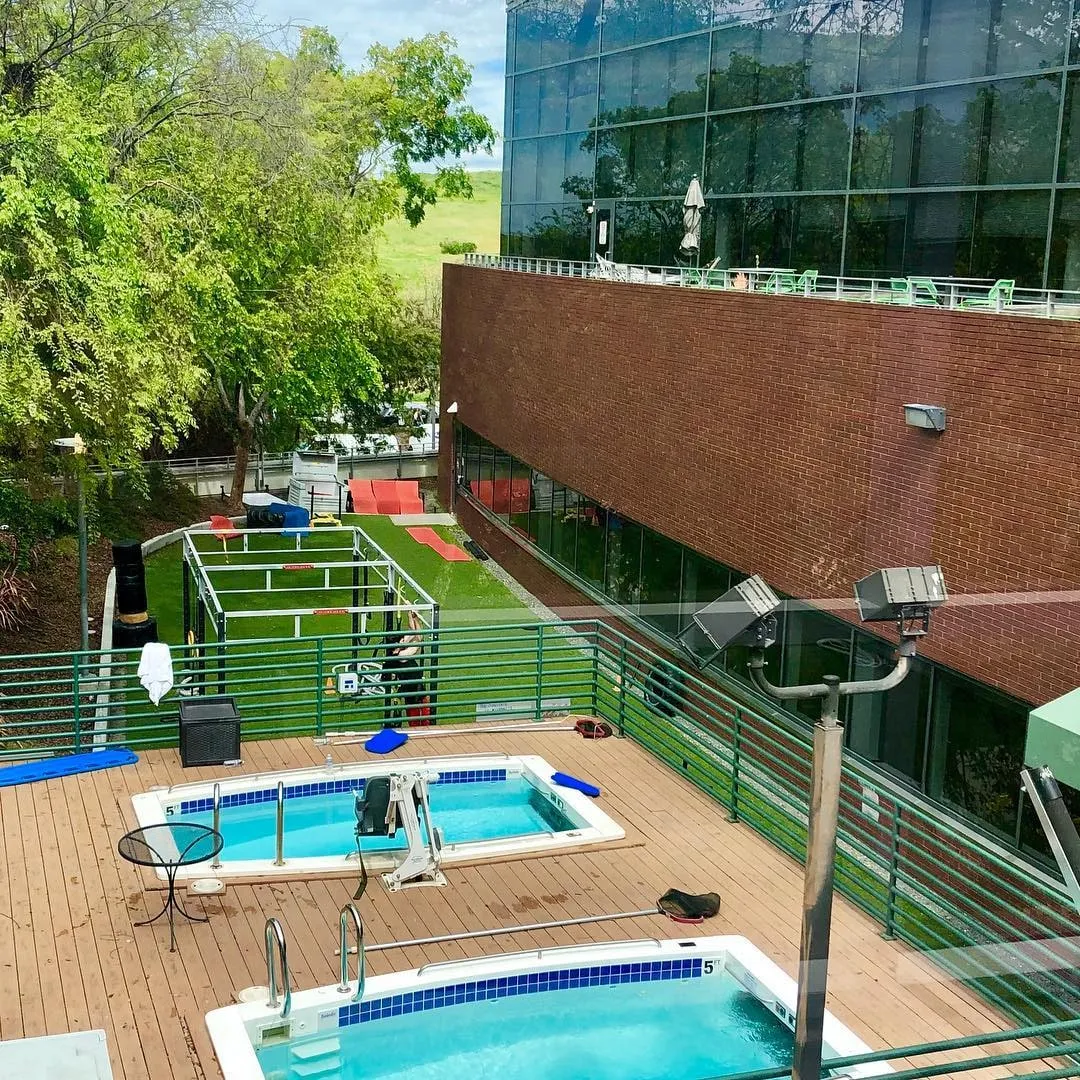 Outdoor wooden deck with two small blue pools and a turf training area beside a brick and glass building