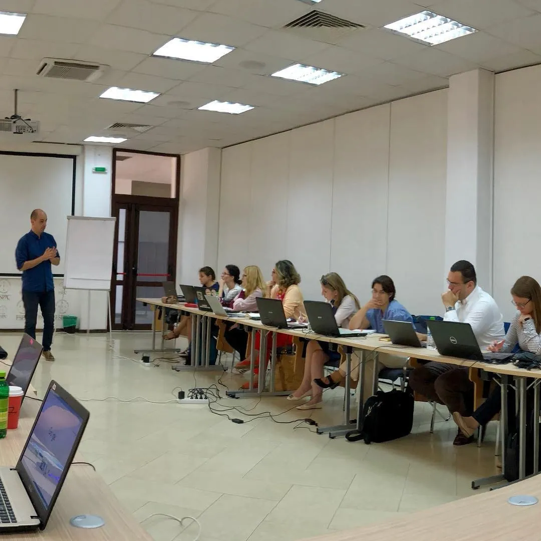 Instructor in a blue shirt presents to a long table of students with laptops in a bright classroom.