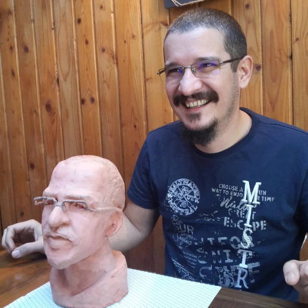 Man with glasses smiling next to a clay bust that looks like him, against a backdrop of wooden planks.