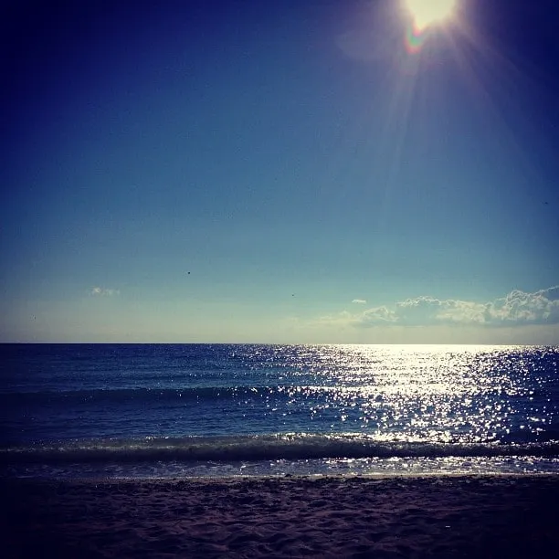 Sunny beach with dark sand, sparkling blue ocean, and a bright sun in a clear sky