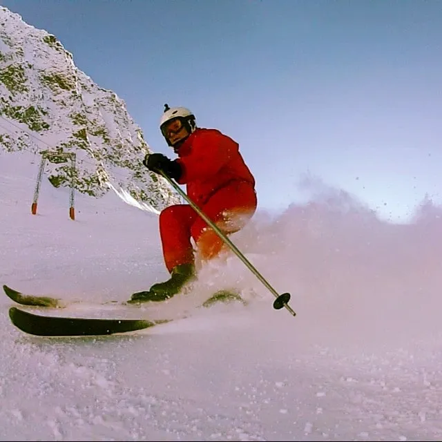 A skier in a red suit and white helmet makes his way down a snowy mountain slope through a spray of powder.