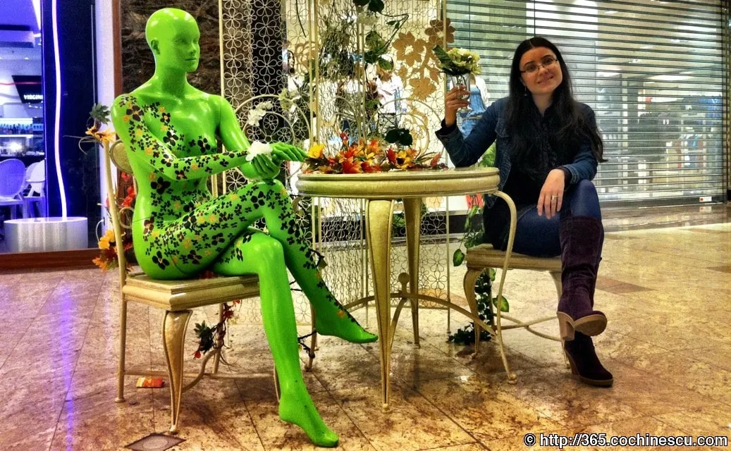 A woman sits at a table next to a green mannequin decorated with flowers.