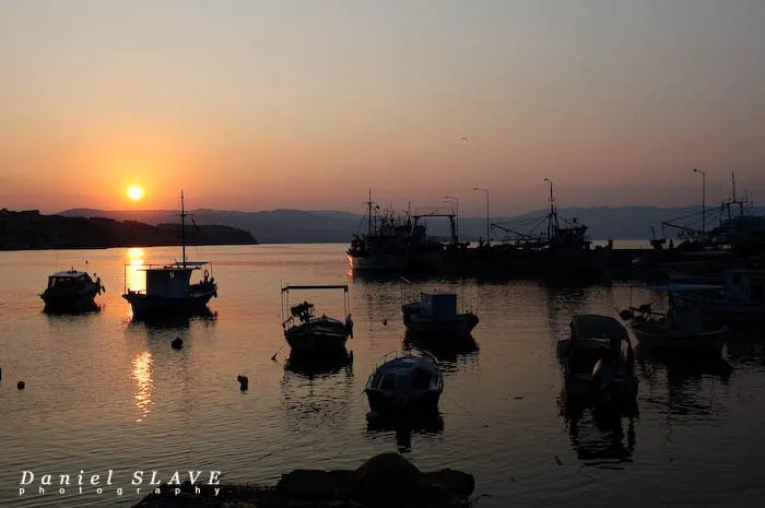 The sun sets over a quiet harbor, amid boats and fishing vessels.