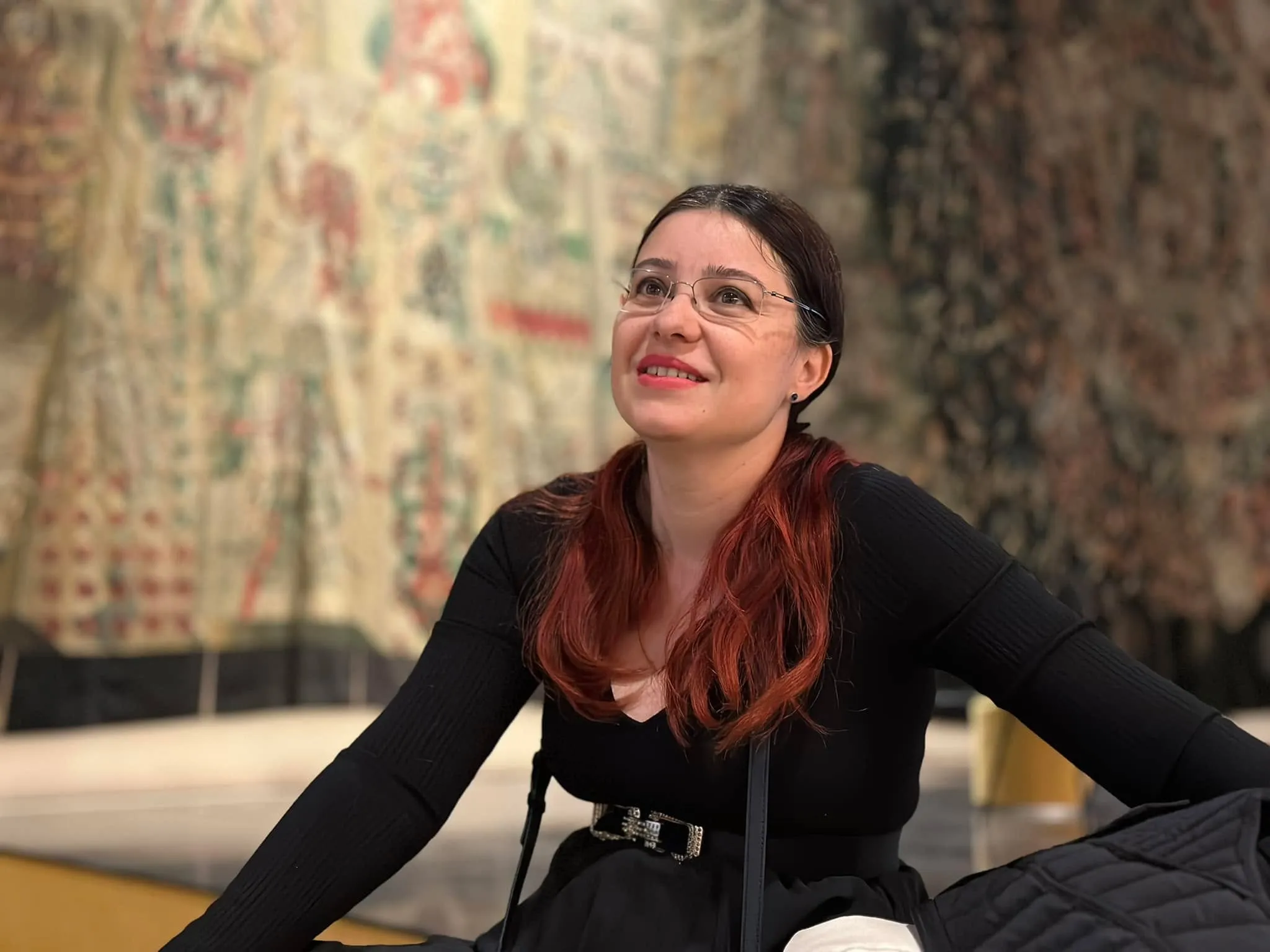 A smiling woman with glasses and reddish-brown hair looks upward while sitting in front of a large embroidered tapestry.