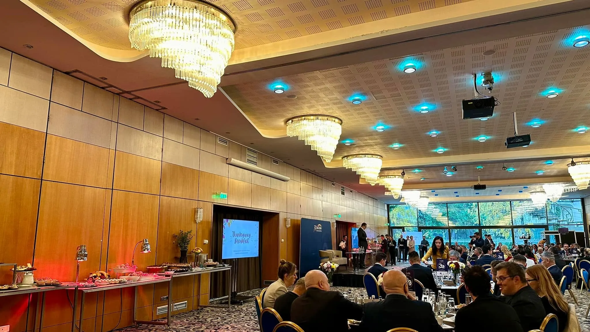 Guests sit at round tables in a chandelier-lit banquet hall with a Thanksgiving Breakfast screen, buffet, and AmCham branding.