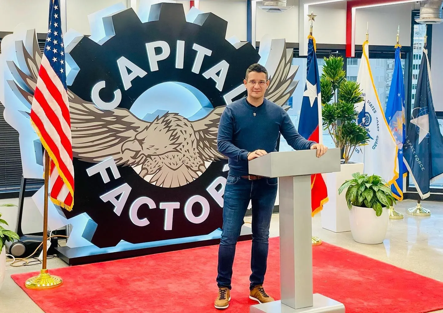 Sebastian Cochinescu stands at a lectern beside the Capital Factory emblem and several flags.