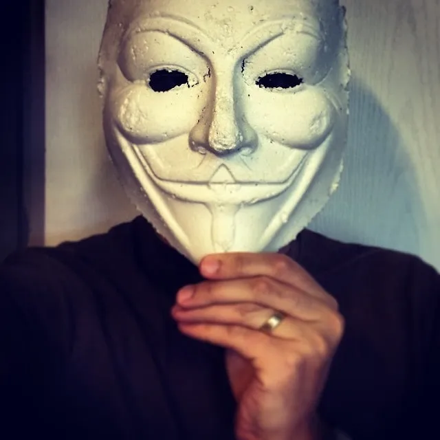 Person taking a selfie while wearing a white Guy Fawkes mask and resting a hand with a ring under the chin.