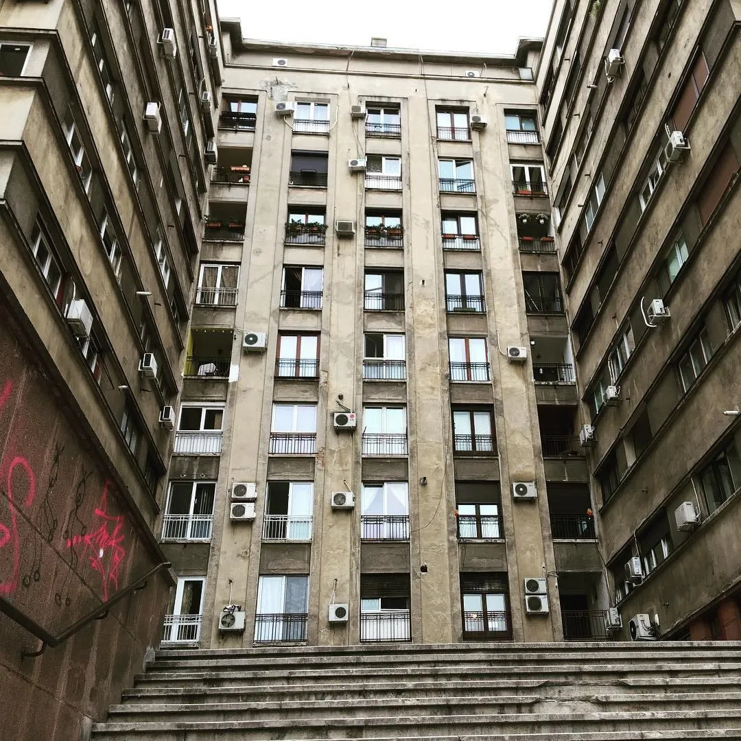 Looking up at a tall beige apartment block with balconies and air conditioners, framed by a courtyard staircase in Bucharest