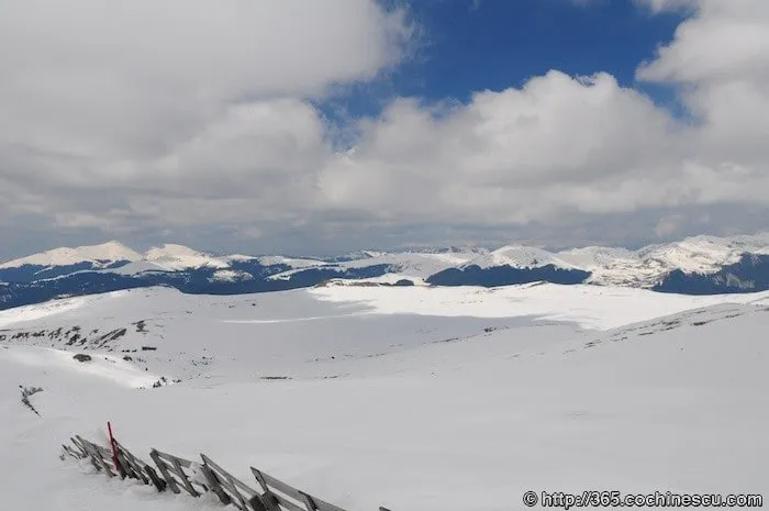 A snow-covered mountain plateau stretches beneath a sky filled with large clouds.