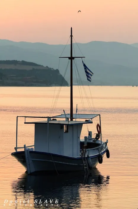 A traditional boat with the Greek flag floats on calm water at sunset.