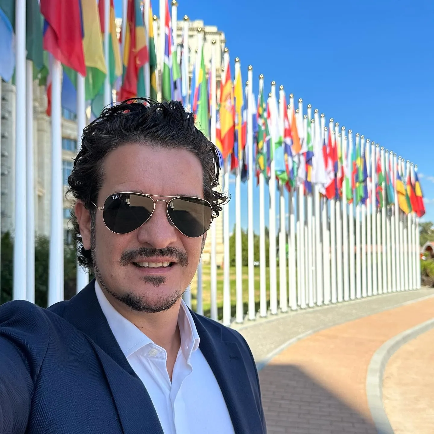 Man in sunglasses and a blue blazer takes a selfie in front of rows of international flags.