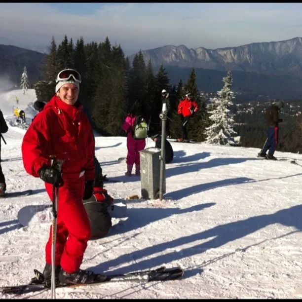Man in a red ski suit on a sunny slope at Clăbucet, with other skiers, pine trees and mountains in the distance.