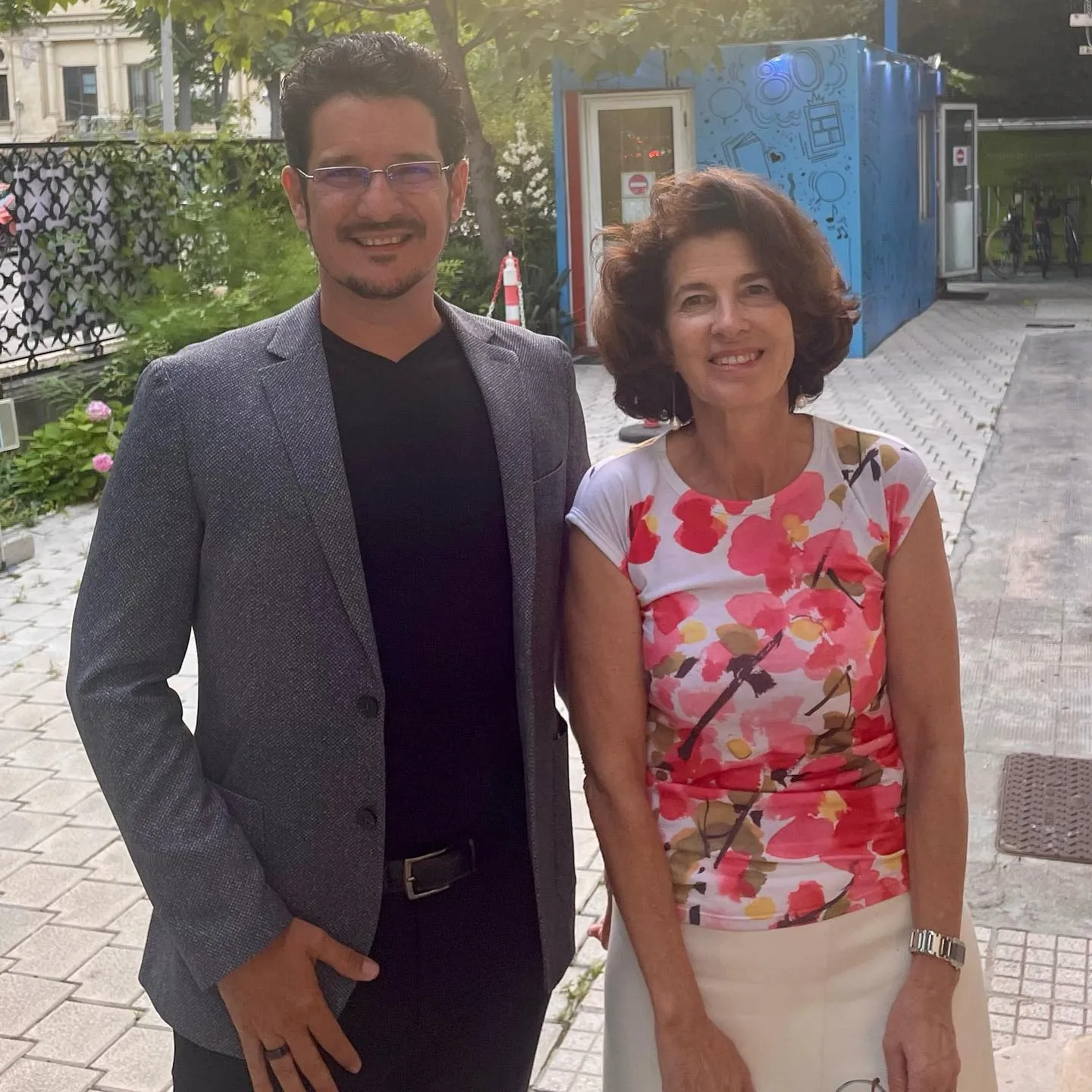 Sebastian Cochinescu and French Ambassador Laurence Auer stand outdoors smiling near greenery and a blue container.