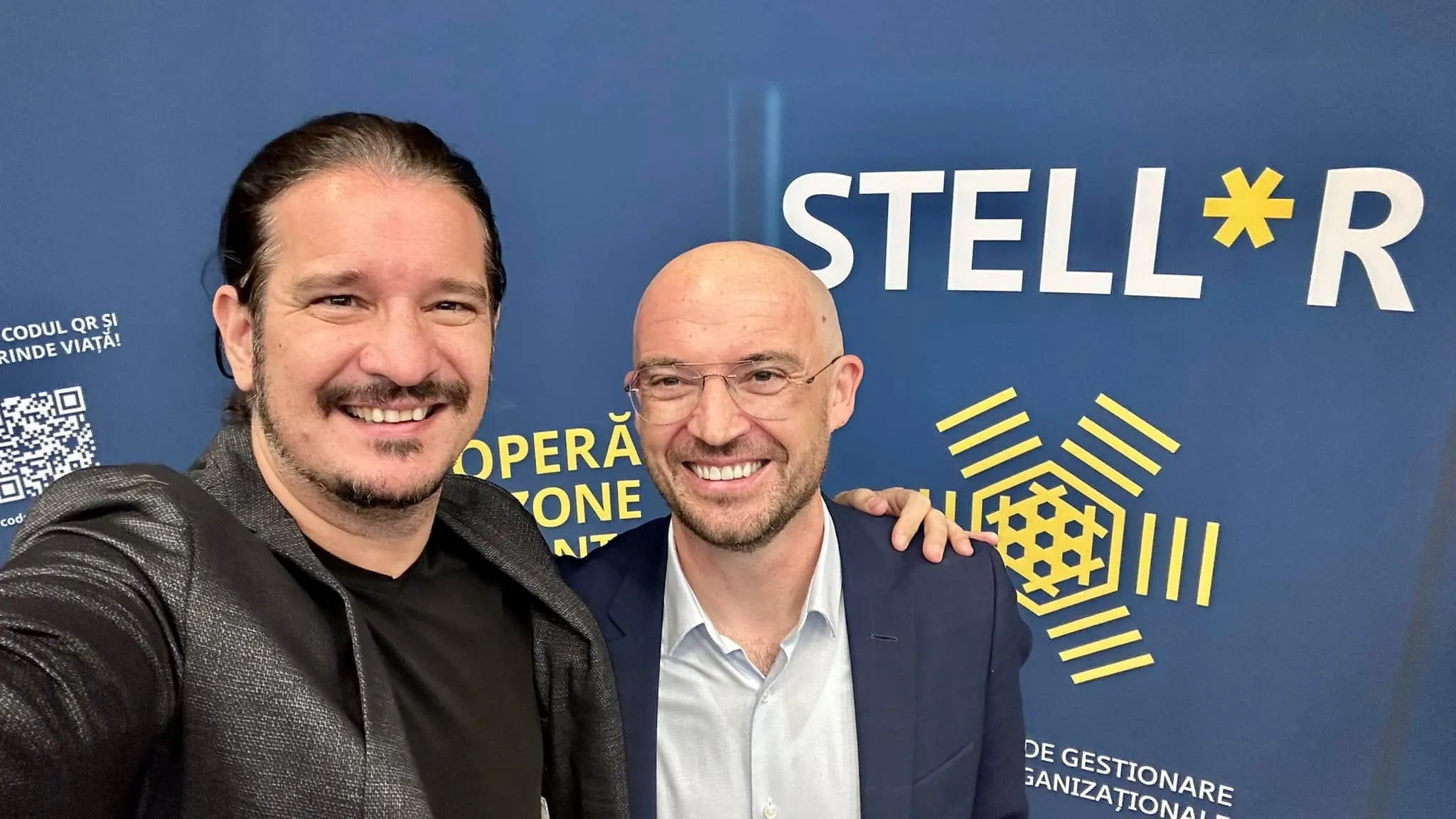 Two smiling men pose for a selfie in front of a blue STELLR backdrop with a gold geometric logo.