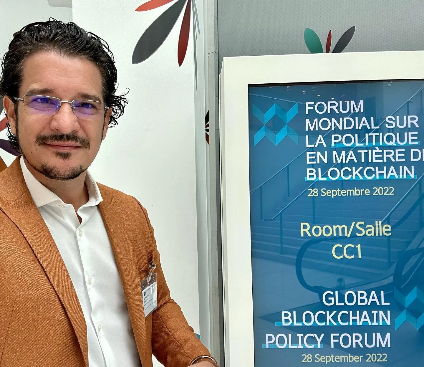 Man in a brown blazer stands beside a Global Blockchain Policy Forum 2022 sign.