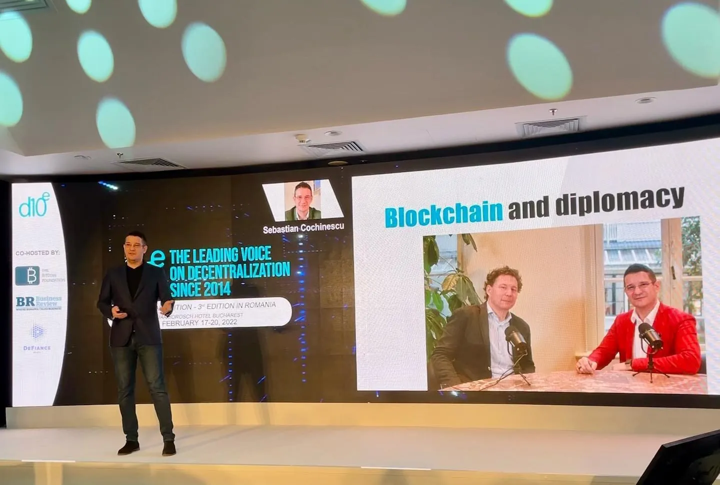Sebastian Cochinescu speaks onstage beside a presentation slide titled Blockchain and diplomacy.