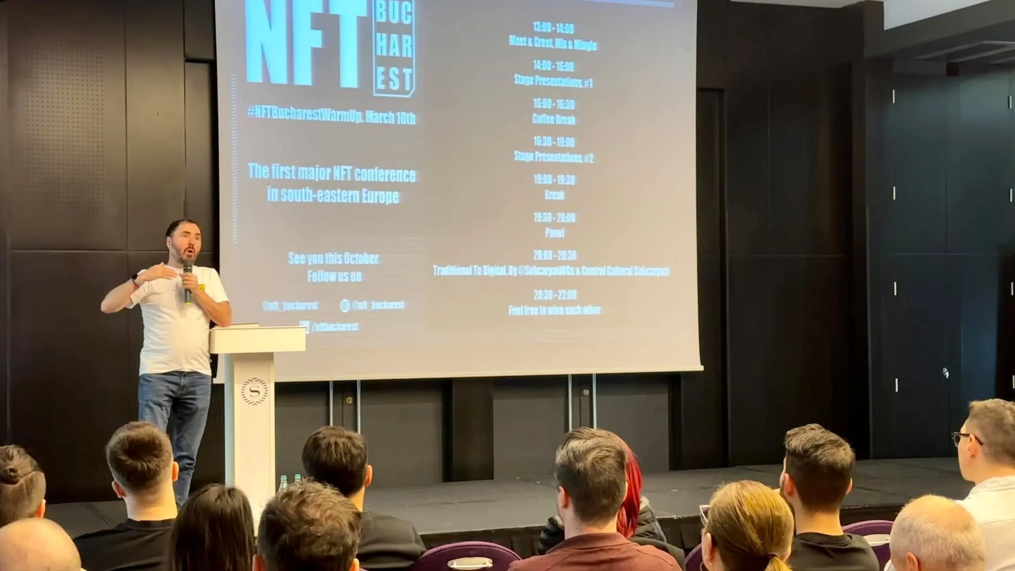 A man in a white t-shirt speaks at a podium beside a large screen for the NFT Bucharest Warm Up event.