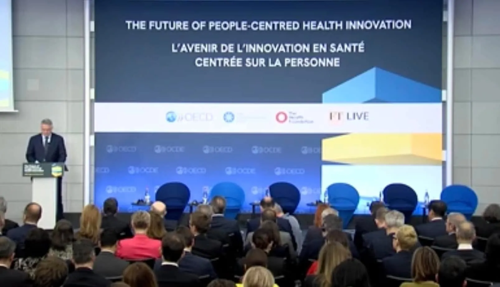 OECD Secretary-General Mathias Cormann speaks at a podium before a large audience and a bilingual health innovation forum screen.