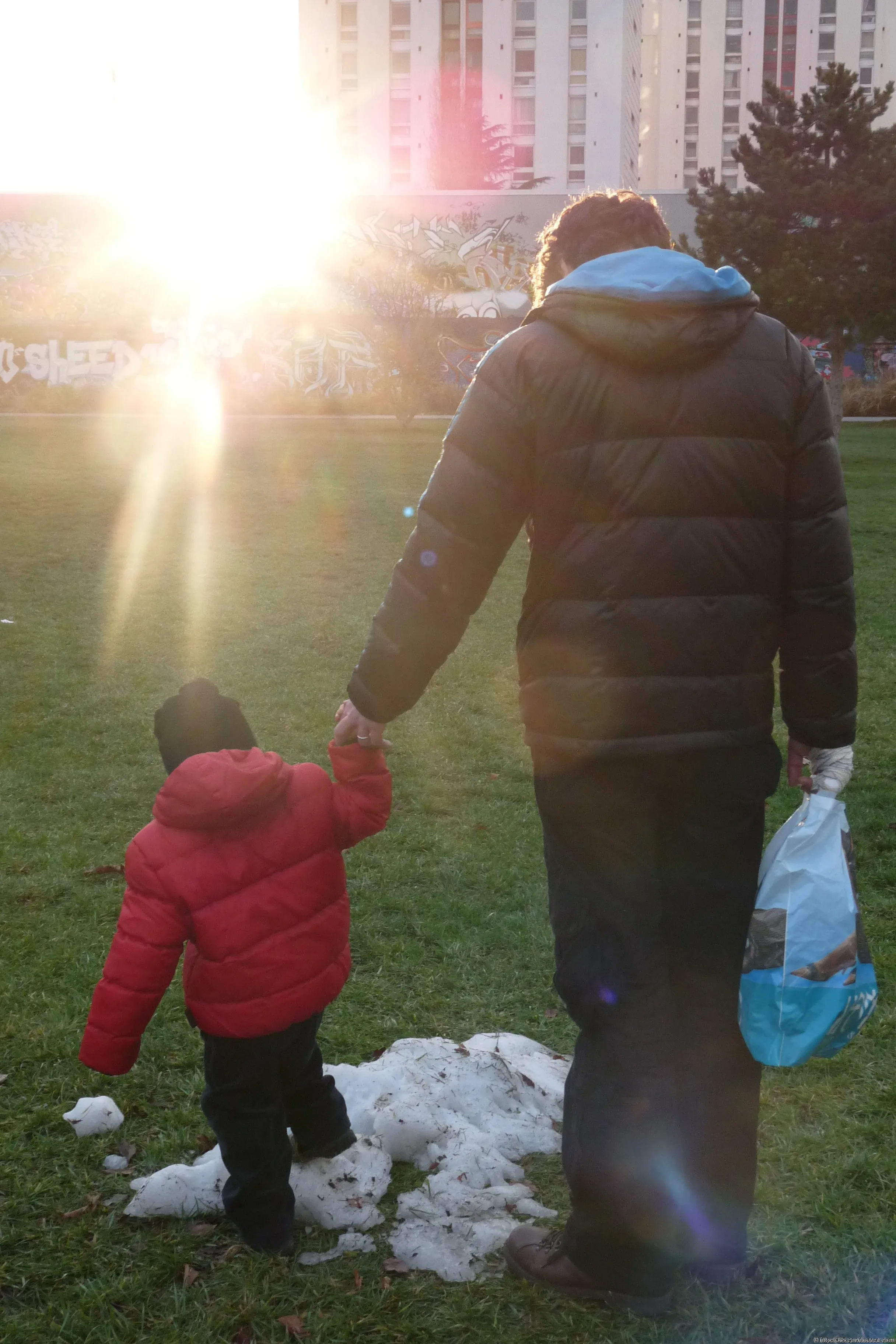 An adult and a child walk hand in hand through a patch of snow in the sunset light.