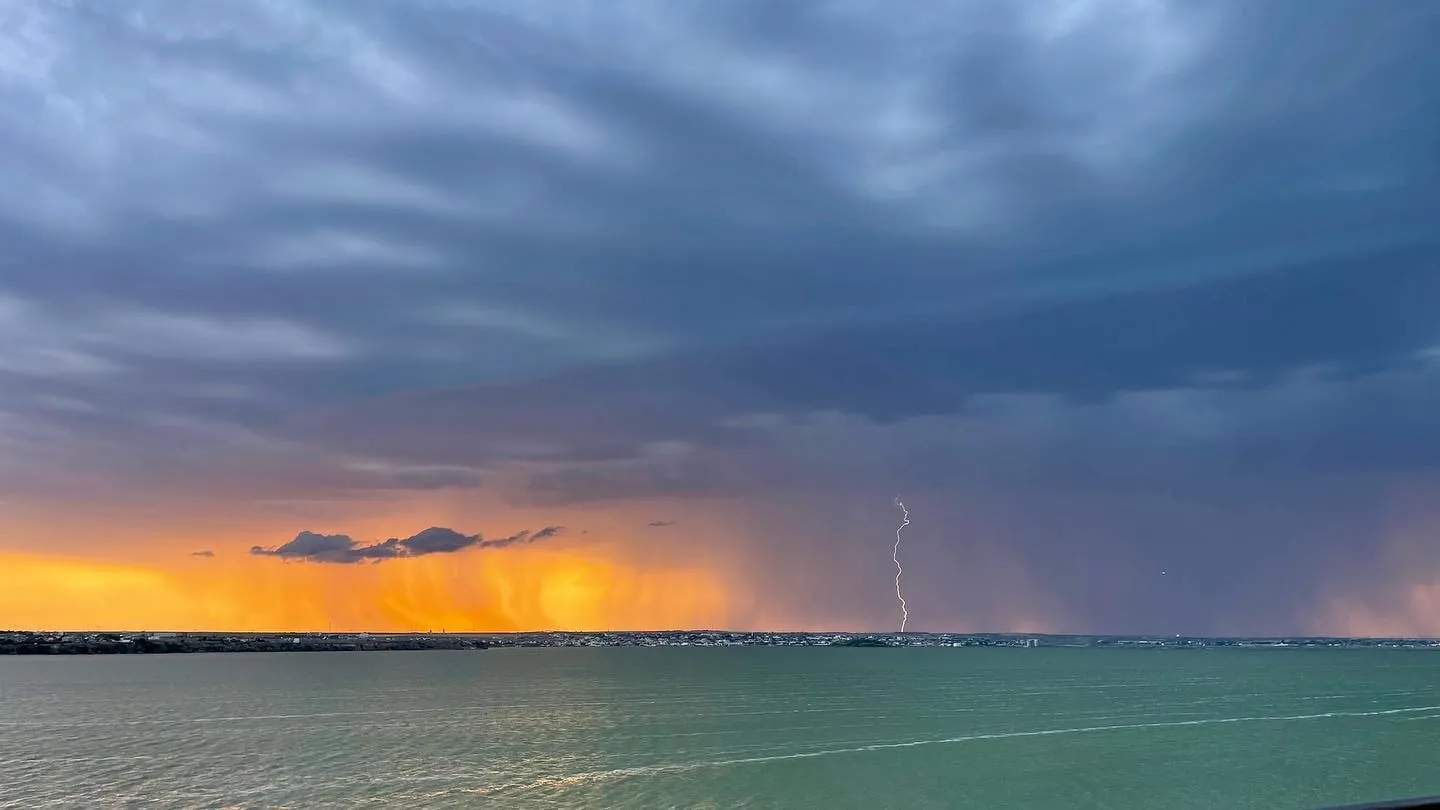 A lightning bolt strikes beneath dark storm clouds as an orange sunset glows across the water.