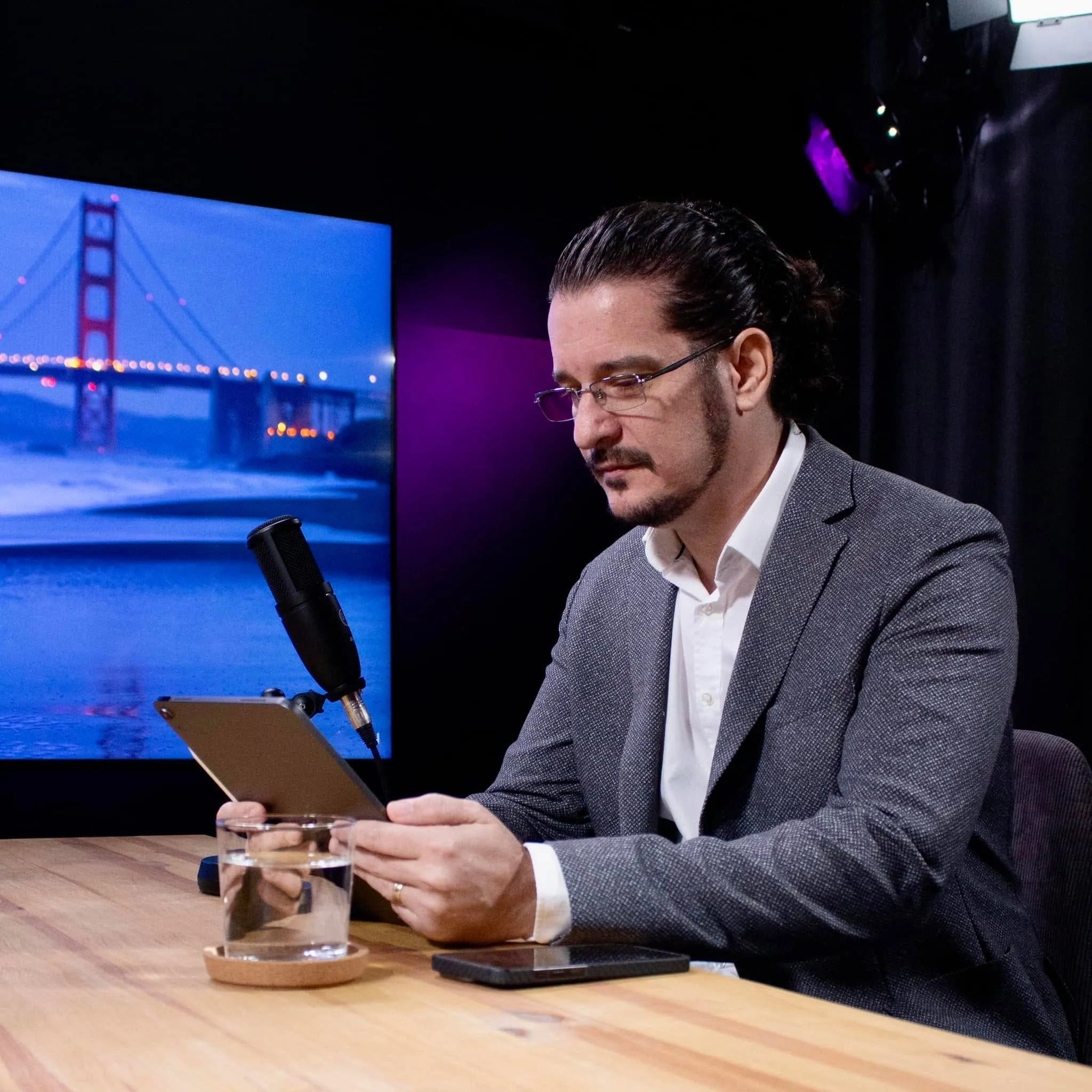 A man in a gray blazer sits at a wooden desk reading a tablet beside a microphone, with a bridge displayed on the screen behind him.