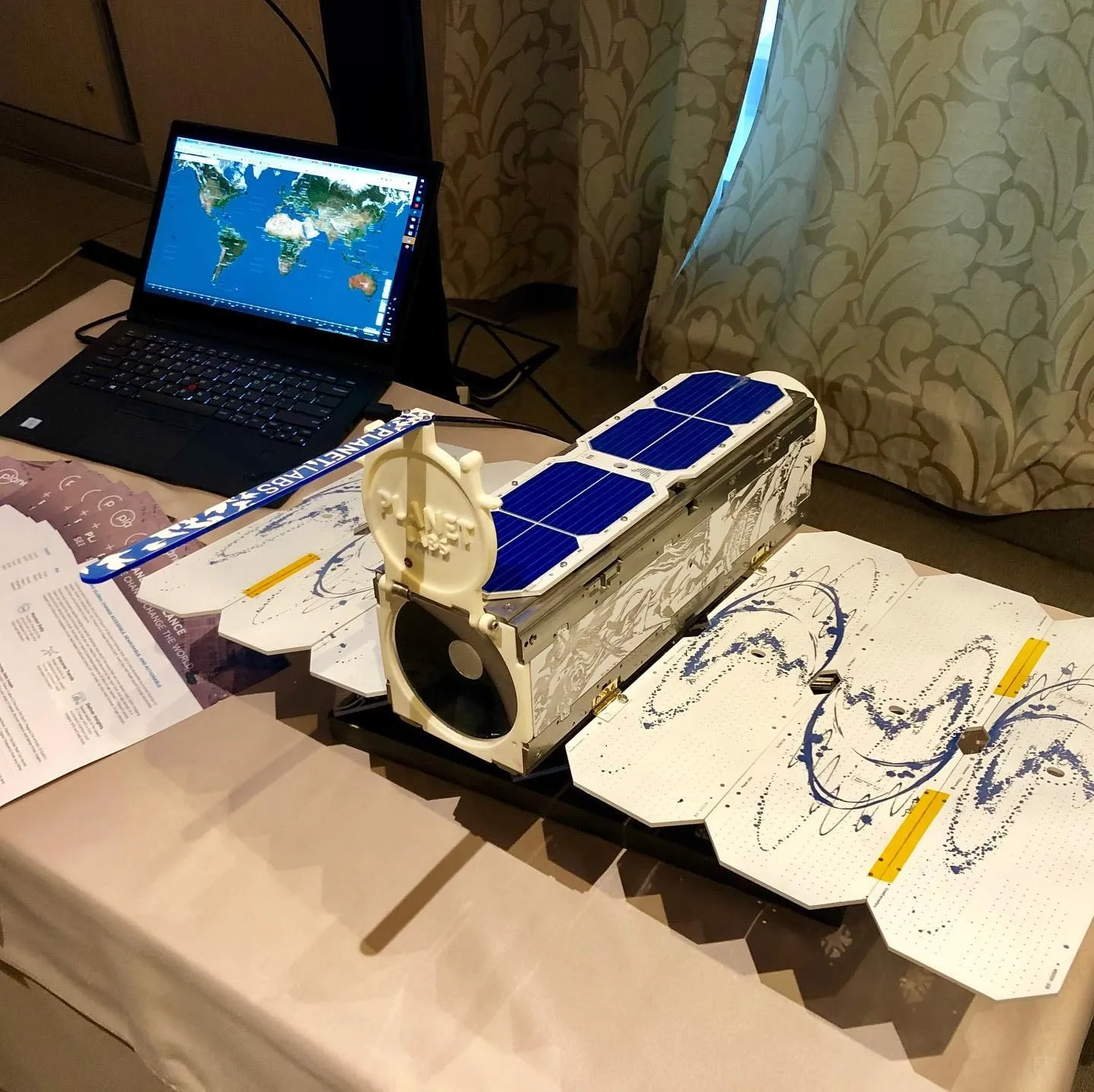 A compact satellite model with solar panels sits beside a laptop displaying a world map.