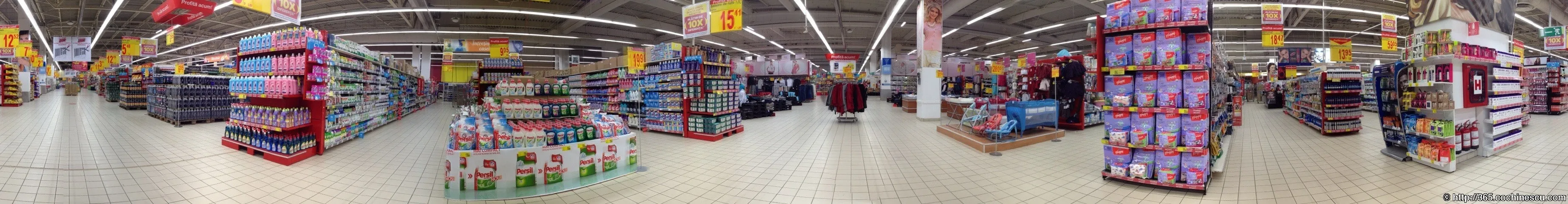 A wide panorama captures the aisles and the numerous yellow signs in a supermarket.