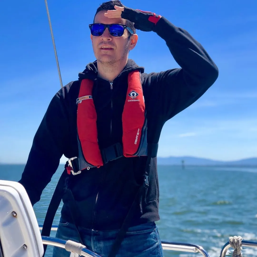Man in a red life vest and sunglasses shields his eyes while standing on a sailboat under a clear blue sky