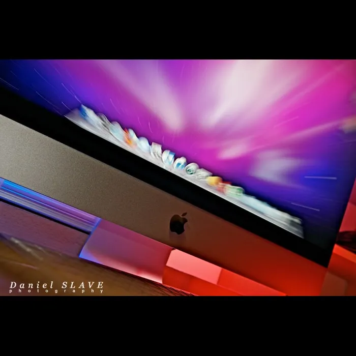 An iMac computer is photographed at an angle, in blue, pink, and red lights.