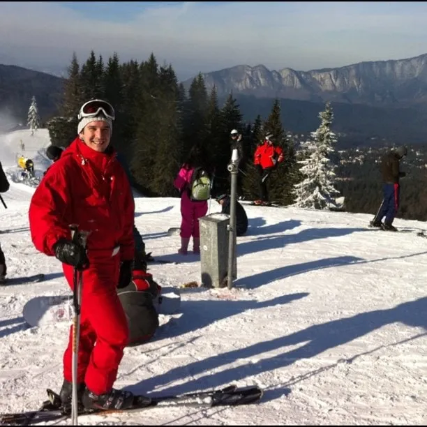 A man in a red ski suit on a slope at Clăbucet, with pine trees, other skiers, and mountains in the background.