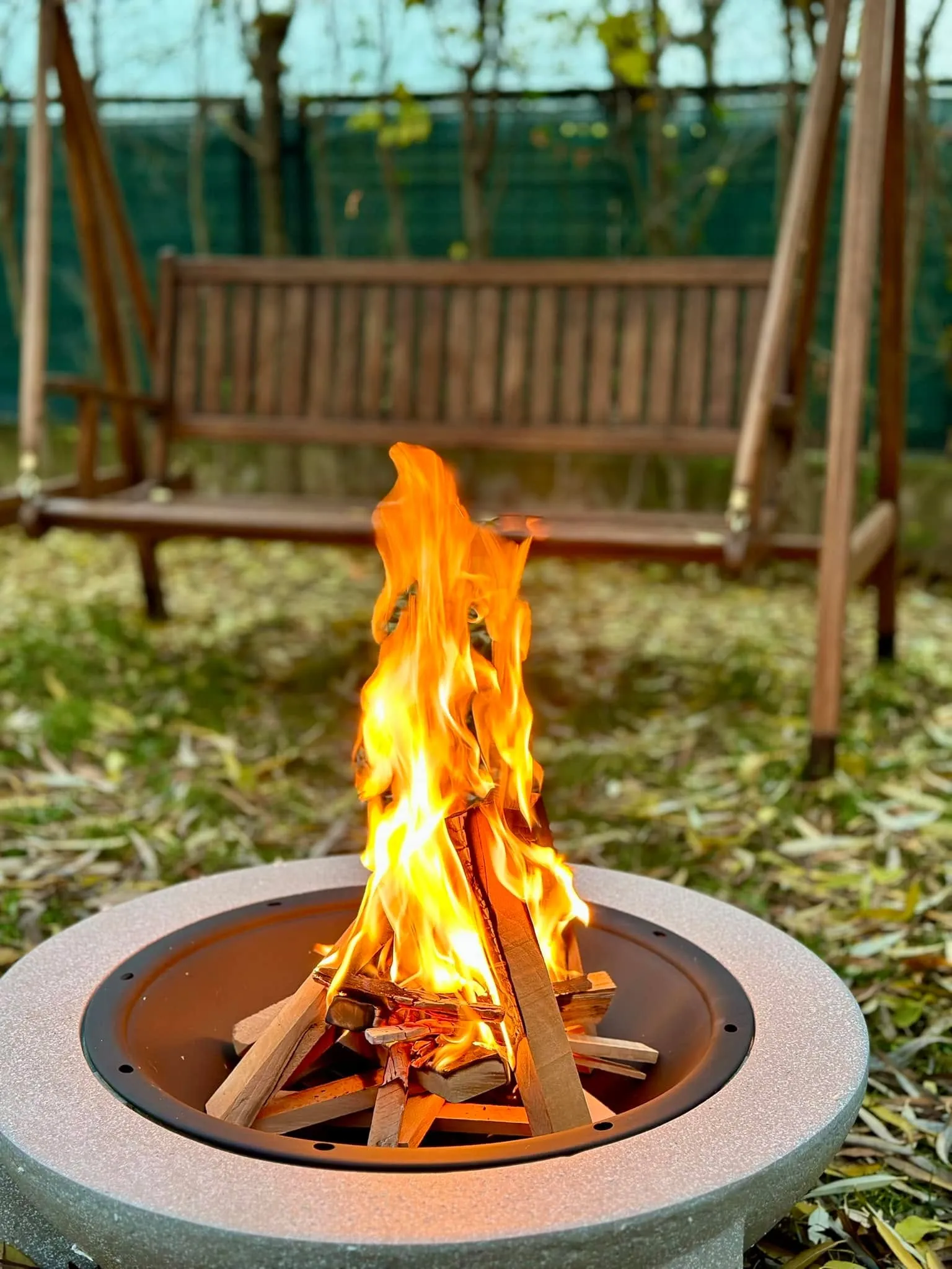 Bright orange flames rise from stacked wood in a round outdoor fire pit, with a wooden porch swing and autumn leaves in the background.