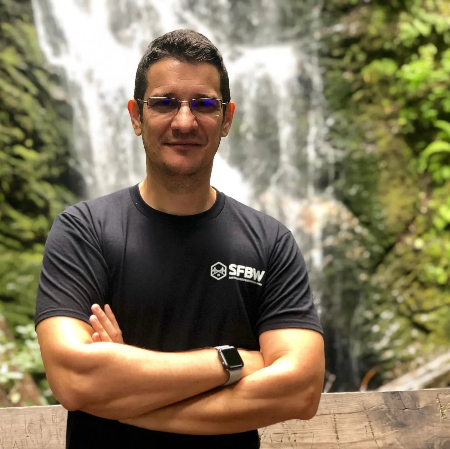 A man in glasses and a black SFBW t-shirt stands arms crossed before a tall waterfall in the forest.