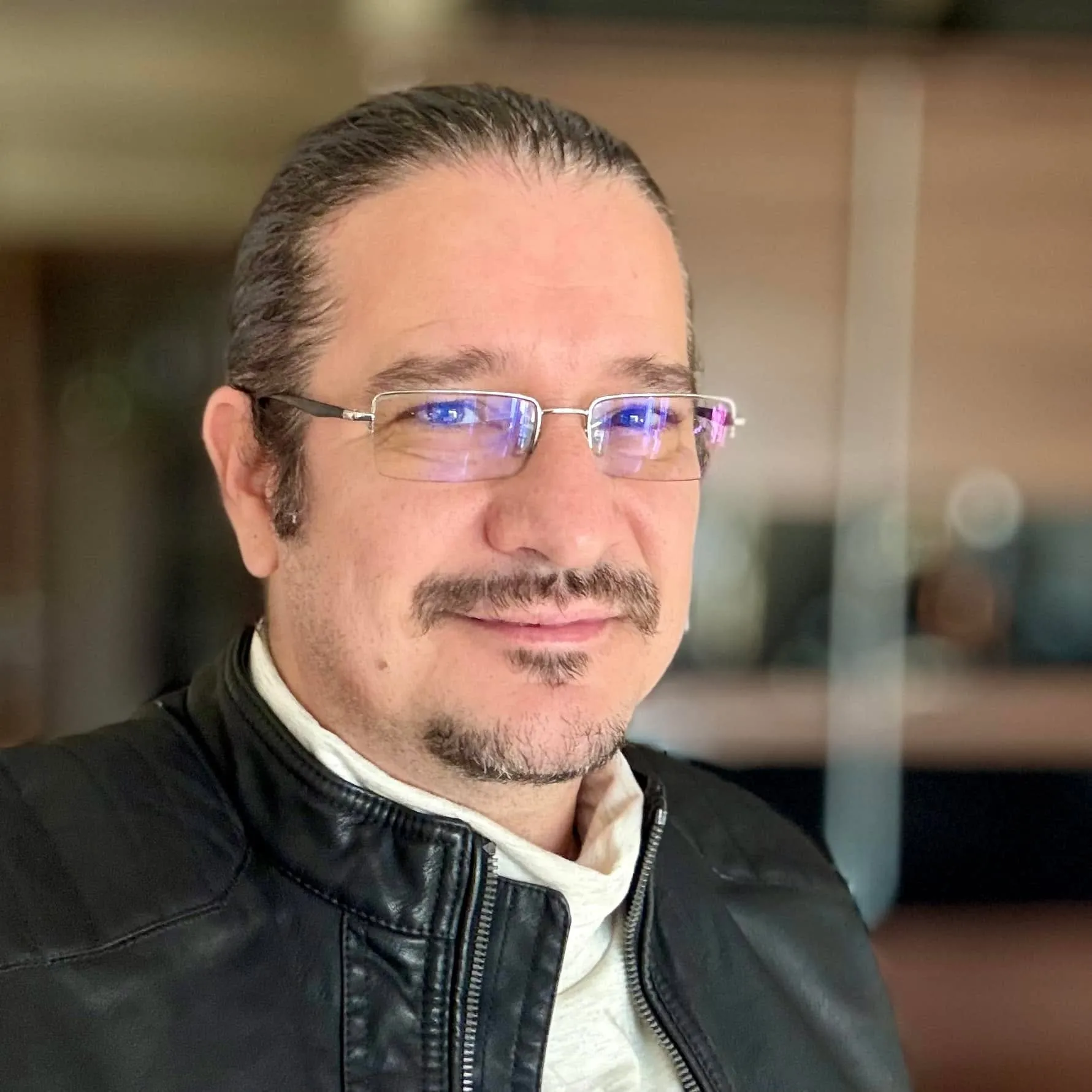 Sebastian Cochinescu smiles for a close-up portrait while wearing glasses and a black leather jacket.