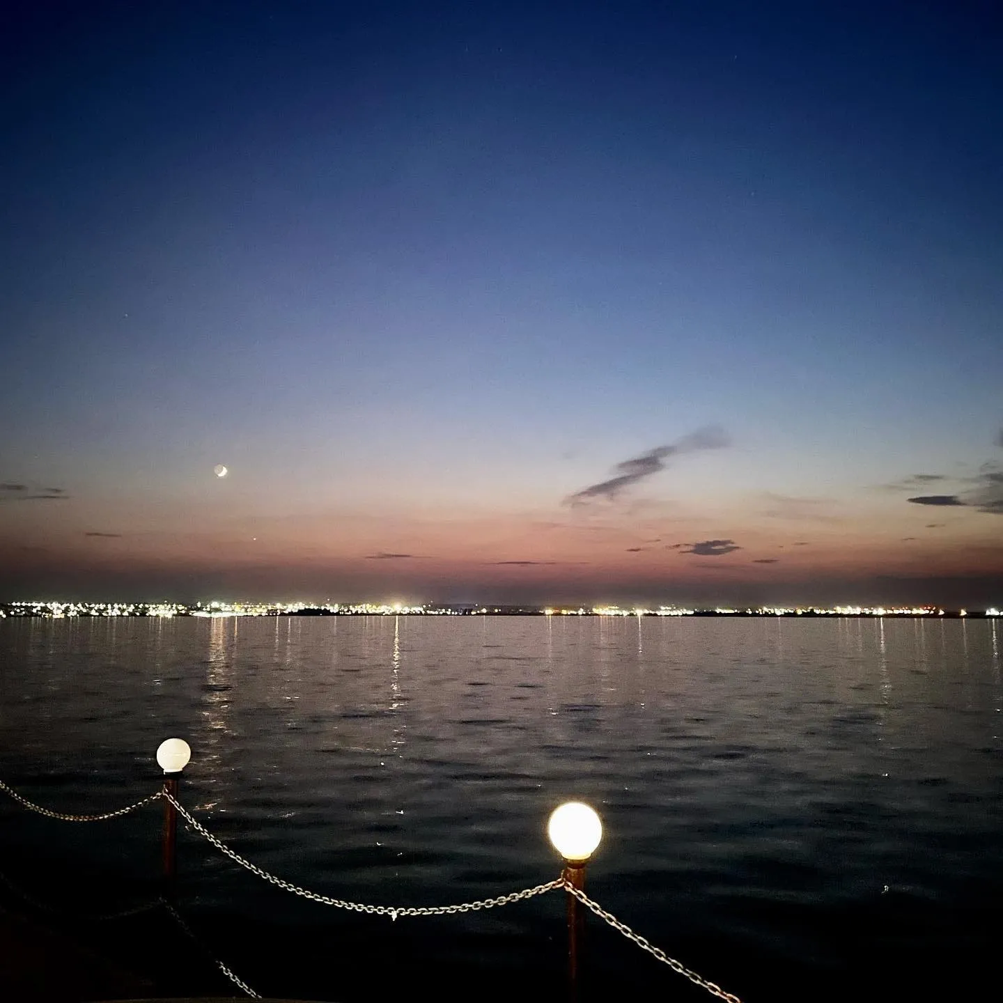 A crescent moon hangs over a calm lake at dusk as distant city lights reflect on the water.