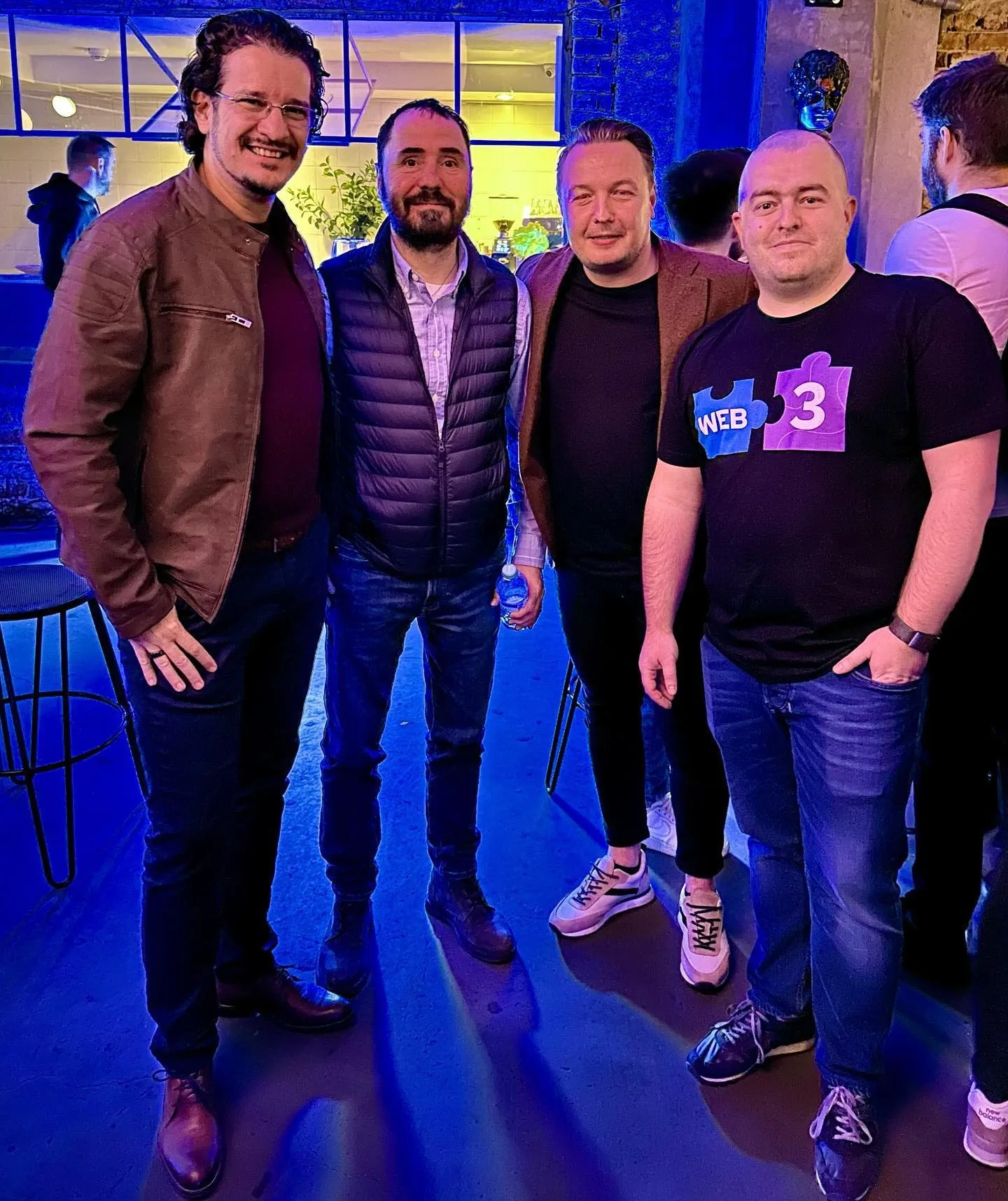 Four men pose together indoors under blue lighting, one wearing a Web3 puzzle-piece shirt.