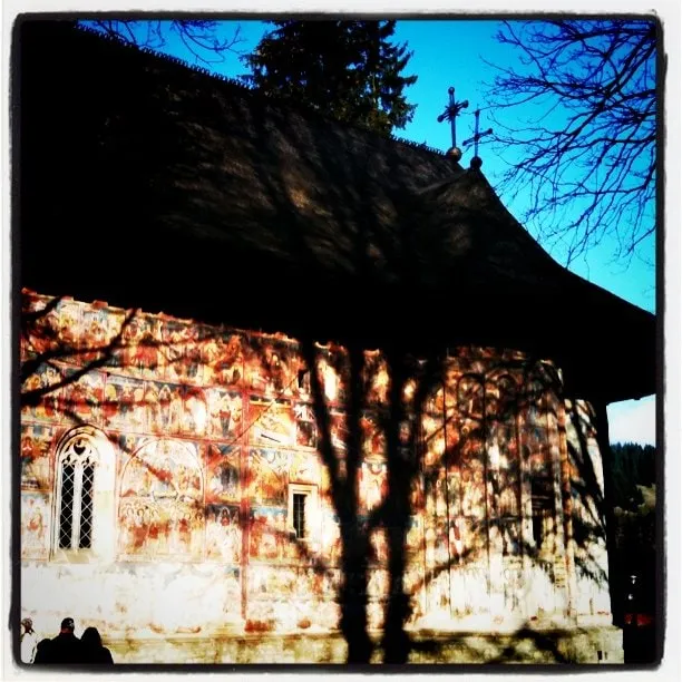 The fresco-covered façade of Humor Monastery, shaded by a tree against a blue sky.