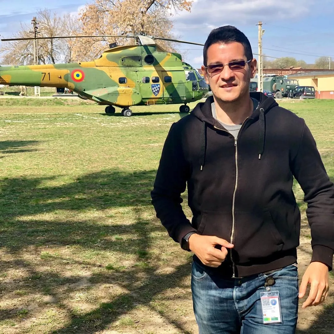 A man in sunglasses and a black hoodie stands on grass in front of a camouflage Romanian military helicopter.