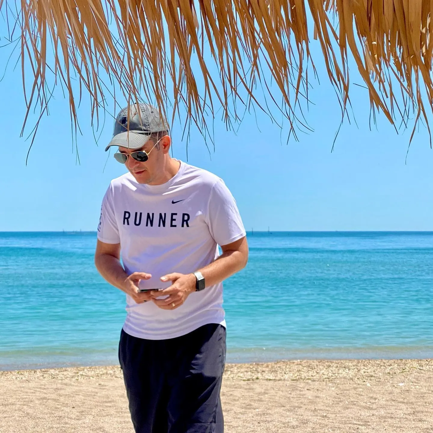 A man wearing sunglasses and a cap stands beneath a thatched beach umbrella beside the turquoise sea.