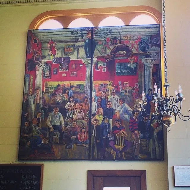 Large mural depicting a café scene full of people, on a wall with arched windows.