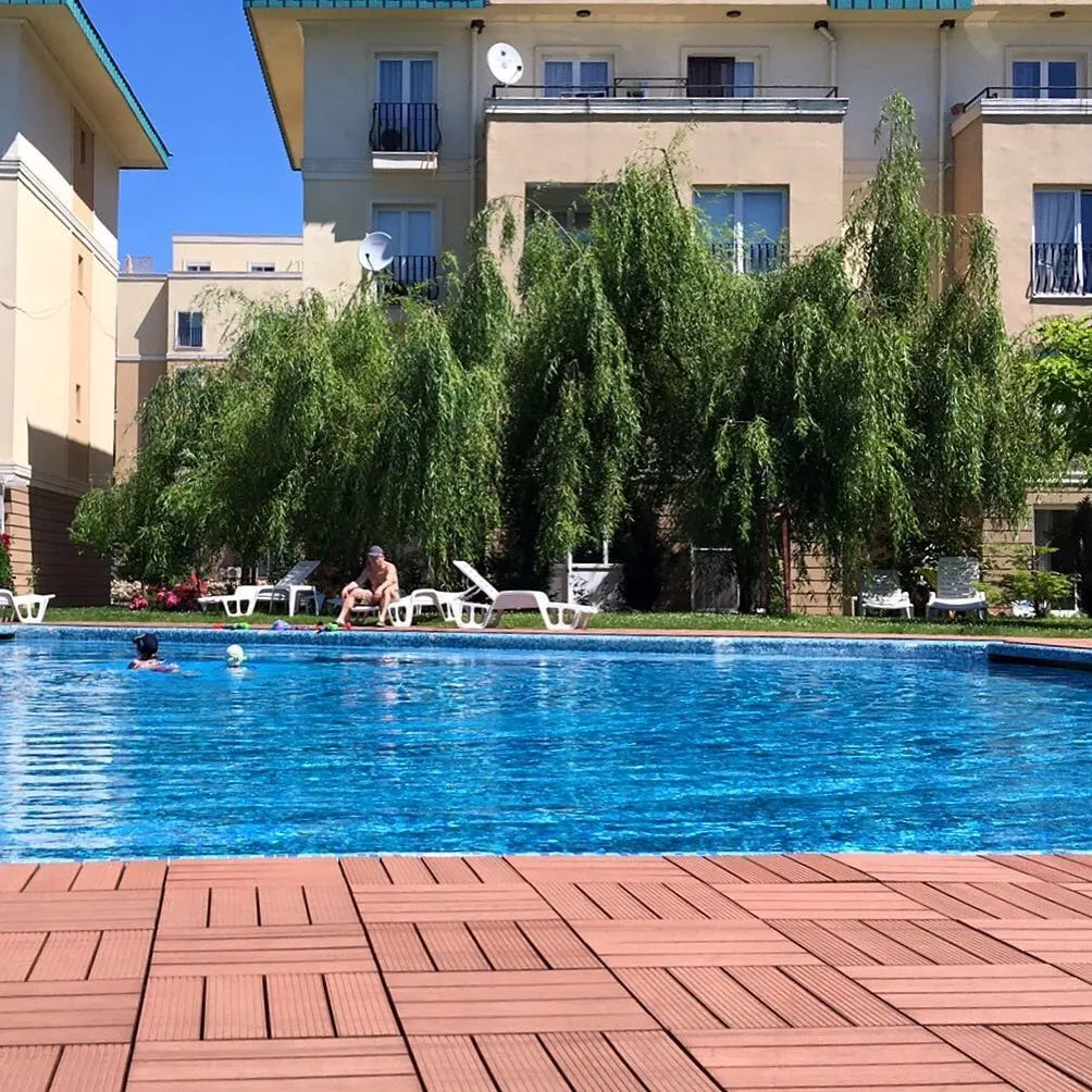 Outdoor pool with people swimming, a man on a lounge chair, and beige apartment buildings behind a willow tree