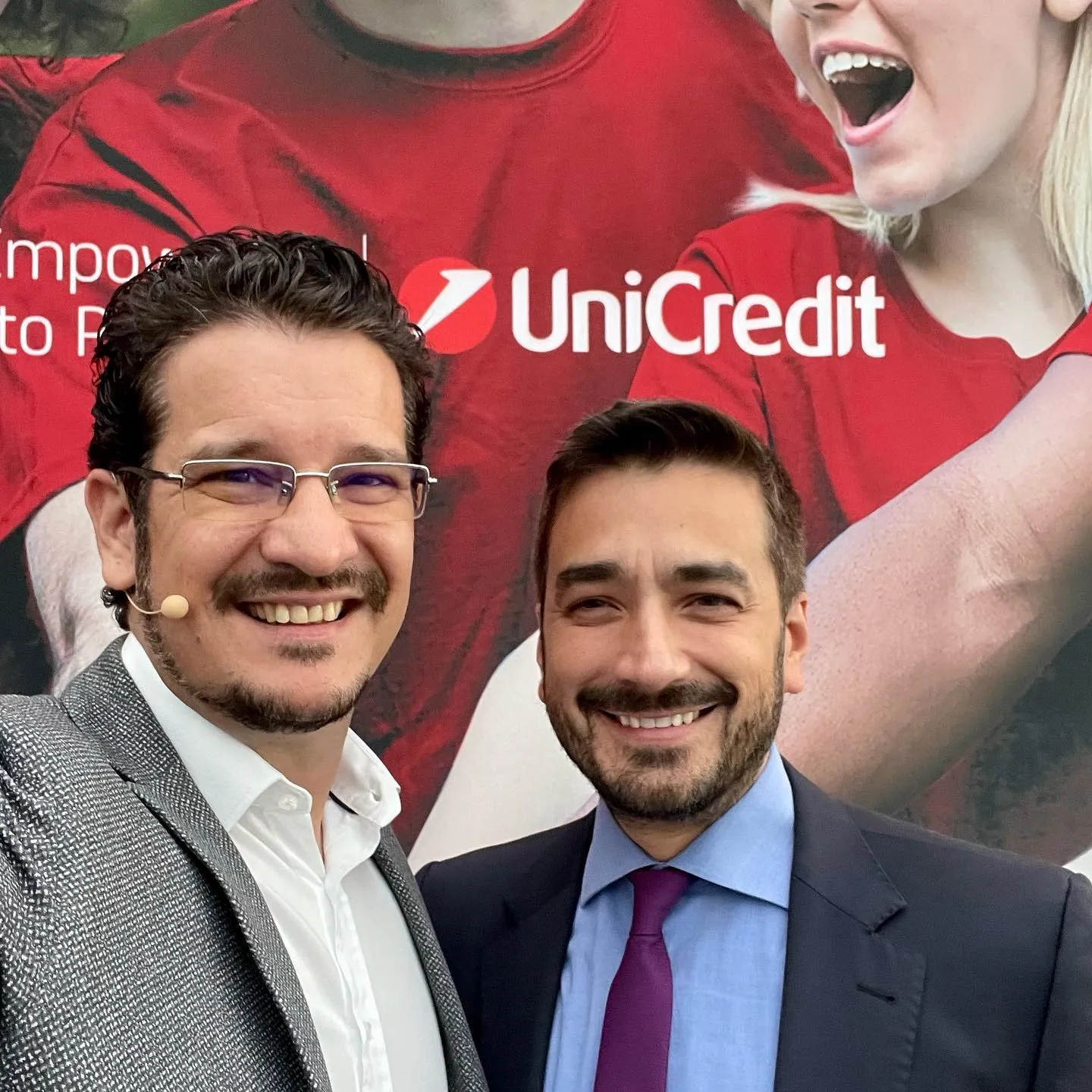 Sebastian Cochinescu and another man smile for a selfie in front of a large UniCredit display.