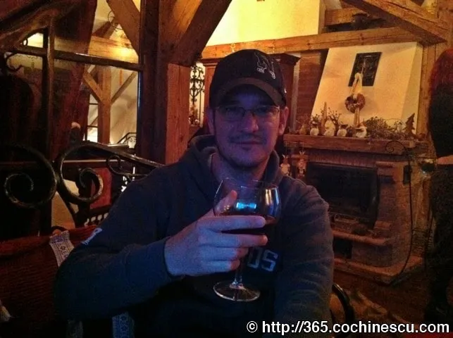 A man with a cap and glasses holds a glass of wine in a rustic interior.